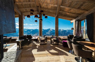 Moderner Loungebereich mit Holzdecke, dunklen Polstermöbeln und Panoramafenstern mit Blick auf verschneite Berge.