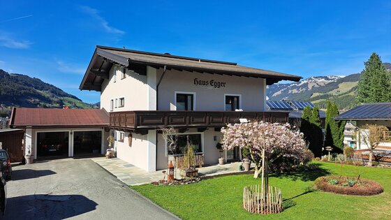 Zweistöckiges Wohnhaus mit Holzbalkon, Garten und Garage vor Bergkulisse.