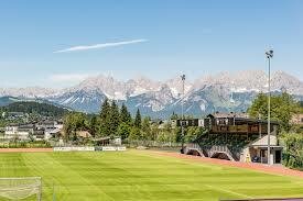 Fußballplatz mit Tribüne vor einem Gebirgspanorama mit schneebedeckten Gipfeln unter blauem Himmel.