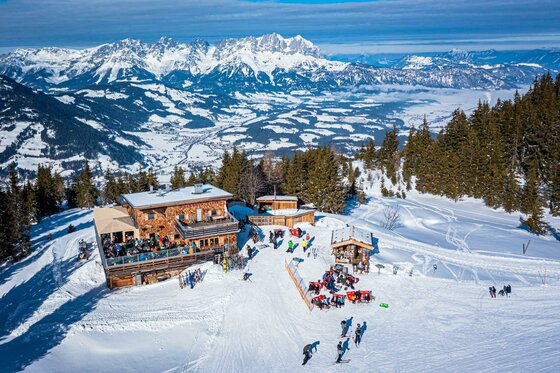 Berghaus Tirol im Winter Berghütte aus Holz im Schnee mit Skifahrern auf der Terrasse vor einem Gebirgspanorama.