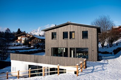 Außenansicht Waldnest Modernes Holzhaus in verschneiter Alpenlandschaft vor blauem Himmel.