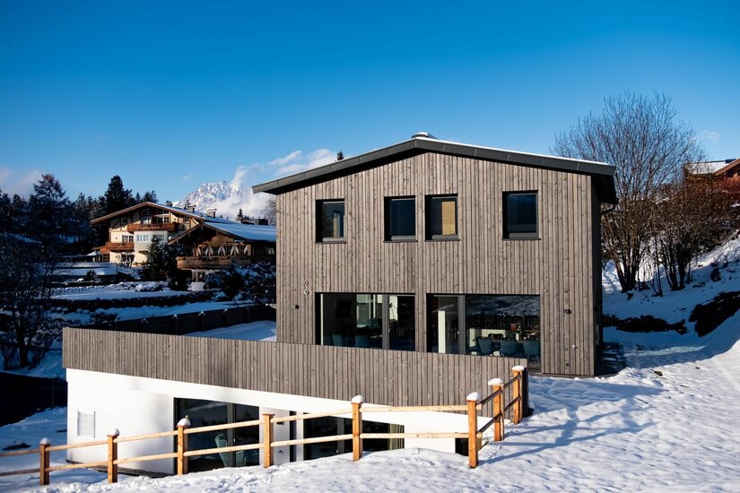 Außenansicht Waldnest Modernes Holzhaus in verschneiter Alpenlandschaft vor blauem Himmel.