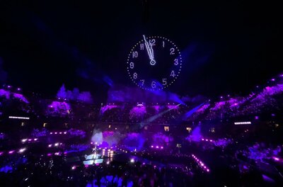 Blick in ein Stadion bei Nacht mit lila Beleuchtung und einer leuchtenden Uhr am Nachthimmel über der Bühne.