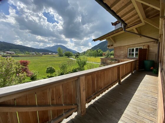Aussicht vom Balkon HB, GB Ein Balkon mit Holzgeländer und Blick auf eine grüne Wiese. Im Hintergrund sind Berge und ein bewölkter Himmel zu sehen.