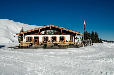 Berghütte aus Holz und Stein mit Terrasse im verschneiten Skigebiet unter blauem Himmel.