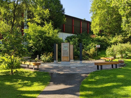 Infoplatz Kitz-Connection Gepflasterter Platz mit hölzernen Infostelen und Sitzbänken inmitten von Bäumen und Rasenflächen.