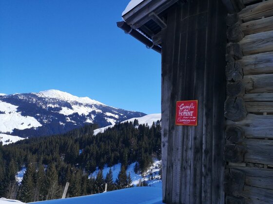 Holzhütte mit rotem Schild vor verschneiter Berglandschaft und Nadelwald unter blauem Himmel.