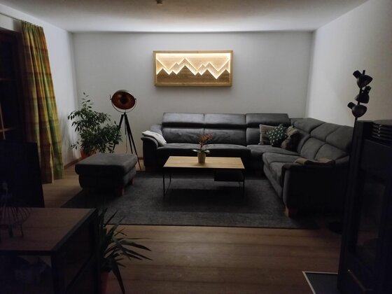 Wohnzimmer mit grauem Ecksofa, Holztisch und beleuchtetem Wandbild in Bergform.