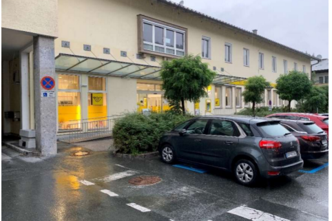 Behindertenparkplatz - Josef-Pirchl-Straße 11 Ein ruhiger Parkplatz mit mehreren Autos vor einem modernen Gebäude. Die Fenster sind beleuchtet und es gibt grüne Pflanzen in der Umgebung.
