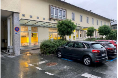 Ein ruhiger Parkplatz mit mehreren Autos vor einem modernen Gebäude. Die Fenster sind beleuchtet und es gibt grüne Pflanzen in der Umgebung.