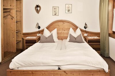 Doppelbett aus hellem Holz mit weißer Bettwäsche, Nachttischen und einem offenen Holzschrank in einem Pensionszimmer.