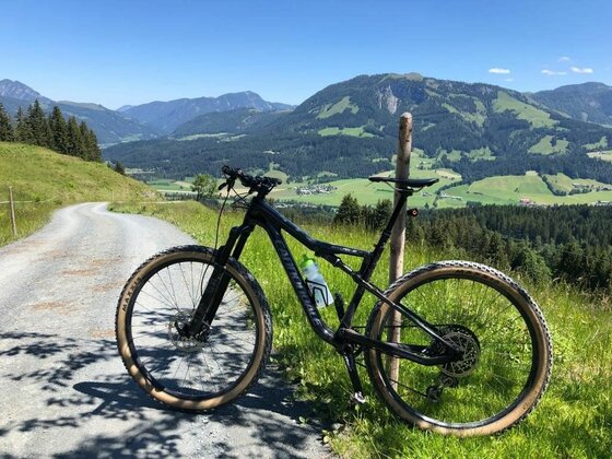 Mountainbike am Harschbichl Ein schwarzes Mountainbike lehnt an einem Holzpfosten neben einem Schotterweg vor einem Bergpanorama.