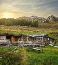 Rustikale Holzhütte mit Terrasse vor bewaldeten Hängen und felsigen Berggipfeln bei tiefstehender Sonne.