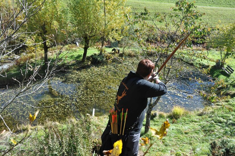 Bogenschießen am Parcours Alte Wacht Person mit Langbogen zielt von einer Anhöhe über einen Teich auf ein Ziel im Wald.