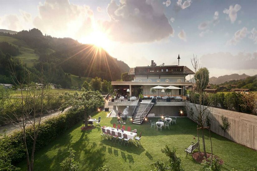 Außenansicht eines Gasthofs mit Gartenrestaurant, Terrasse und angrenzender Wiese vor bewaldeten Bergen.