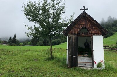 Lindenthal Kapelle Eine kleine Kapelle steht in einer grünen Wiese, umgeben von Bäumen. Der Himmel ist bewölkt und die Landschaft wirkt ruhig und idyllisch.