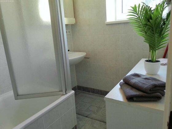 Ein helles Badezimmer mit einer Dusche, einem Waschbecken und einem kleinen Pflanze. Auf der Ablage liegen graue Handtücher und eine weiße Vase.