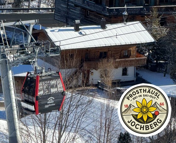 Eine Seilbahn fährt über ein schneebedecktes Gebiet mit einem Holzhaus im Hintergrund. Das Bild zeigt die Umgebung von Jochberg im Winter.
