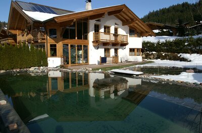 Alpines Holzhaus mit Solaranlage und Schwimmteich im Winter.