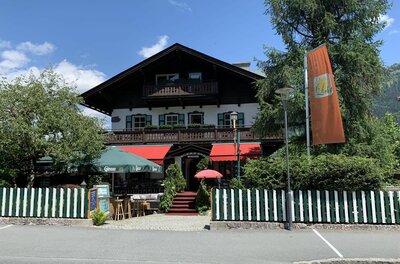 Villa Licht Außenansicht Alpines Gebäude mit Holzbalkonen, grün-weißem Zaun und Außengastronomie unter Sonnenschirmen.