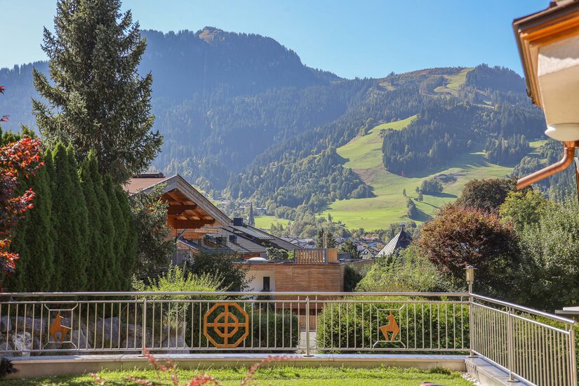 Ausblick Hahnenkamm Streif Blick von einer Terrasse mit Metallgeländer auf bewaldete Berge und Almwiesen unter blauem Himmel.