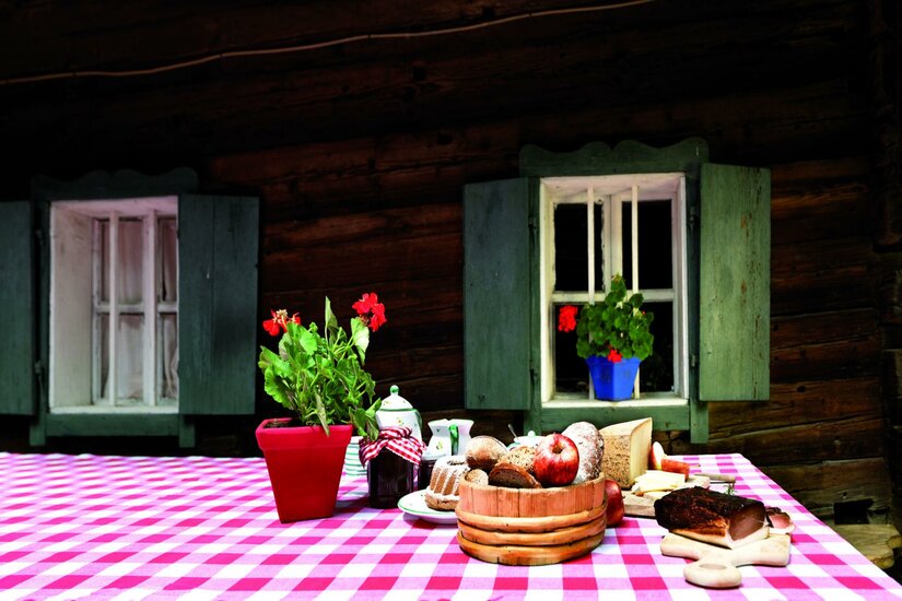 Ein Tisch mit kariertem Tischdeckchen, dekoriert mit frischen Blumen und verschiedenen Lebensmitteln. Im Hintergrund befinden sich zwei Fenster einer Holzhütte.