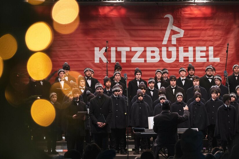 Ein Knabenchor in dunkler Kleidung und Mützen singt auf einer Bühne vor einem roten Kitzbühel-Banner.