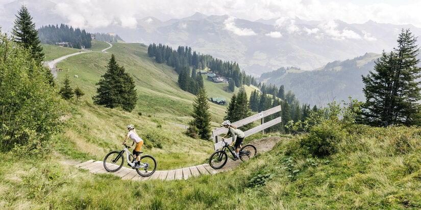 Auf den 360 Flow Trails in Kitzbühel fahren zwei Radfahrer über kurvenreichen Holzweg durch grüne Berglandschaft mit bewaldeten Hügeln.