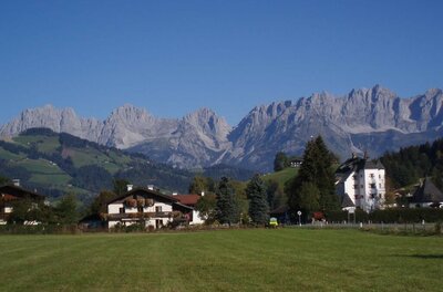 Blick über eine grüne Wiese auf das Haus Zierl und ein weißes Schloss vor dem Gebirgsmassiv Wilder Kaiser.