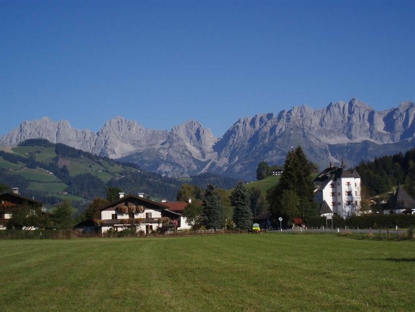 Blick über eine grüne Wiese auf das Haus Zierl und ein weißes Schloss vor dem Gebirgsmassiv Wilder Kaiser.
