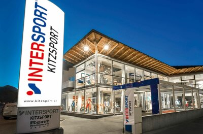 Beleuchtetes Sportgeschäft Intersport Kitzsport bei Dämmerung mit großer Werbesäule im Vordergrund.