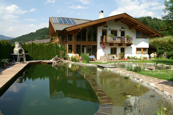 Alpines Wohnhaus mit großem Schwimmteich im Garten und Holzterrasse unter blauem Himmel.