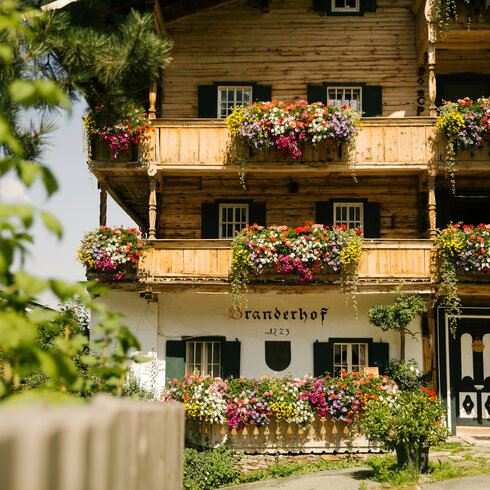 Tradtition Brauchtum Bauernhaus Blumen Details (c) Kitzbuehel Tourismus (1) Ein traditionelles, blumenreiches Haus mit Holzverkleidung, das in einer idyllischen Landschaft steht und Ruhe ausstrahlt.