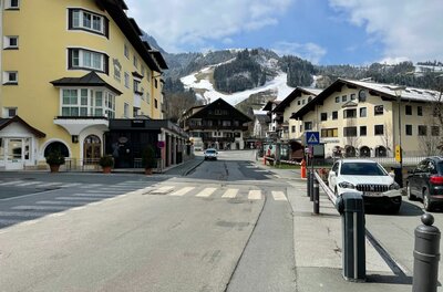 Blick auf eine asphaltierte Dorfstraße mit mehrstöckigen Gebäuden und schneebedeckten Bergen im Hintergrund.