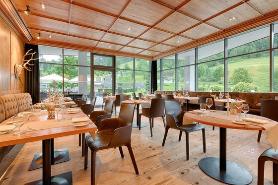 Moderner Restaurantraum mit Holztischen, Lederstühlen und großen Glasfronten mit Blick ins Grüne.