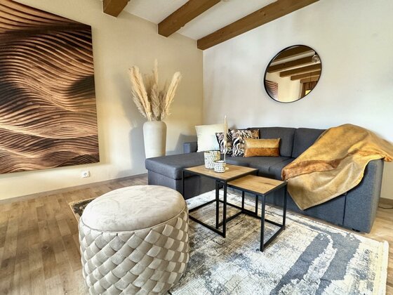 IMG_5272 Large Ein modernes Wohnzimmer mit einer gemütlichen Couch und dekorativen Kissen. Ein stilvoller Tisch und ein runder Pouf ergänzen den Raum, der mit einer Kunstwand und Pflanzen dekoriert ist.