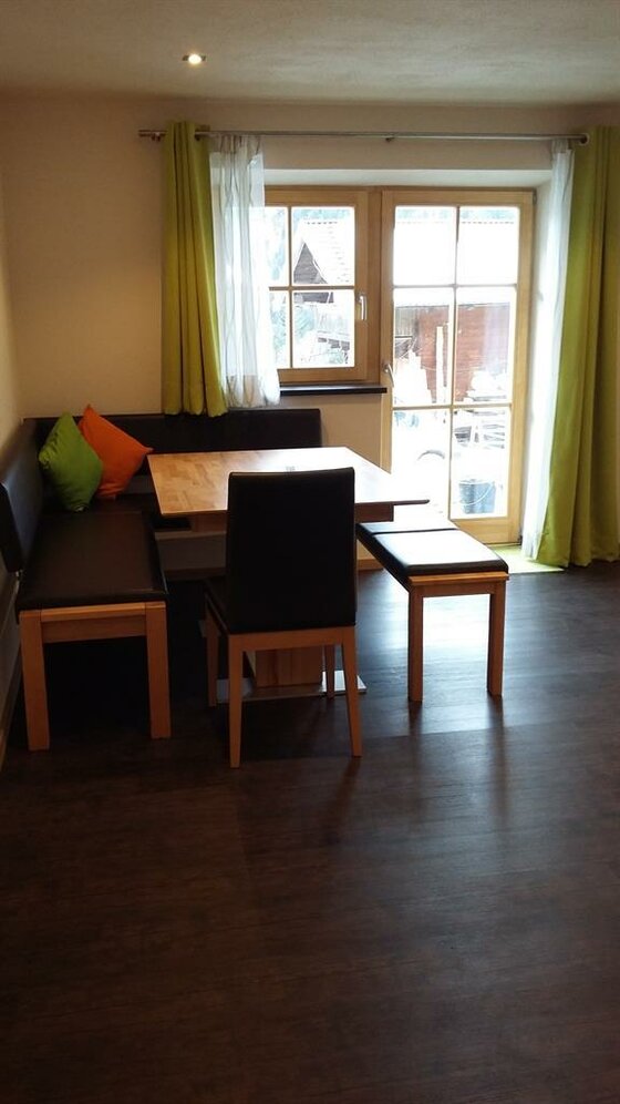 Sitzecke mit Holztisch, Eckbank und Stuhl vor einem Fenster mit grünen Vorhängen.