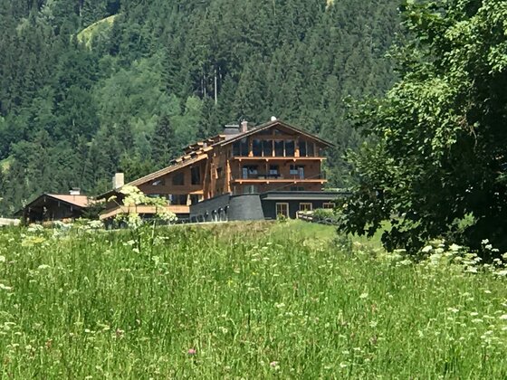 Das Kitz Jochberg Mehrstöckiges Holzgebäude mit Balkonen vor einem bewaldeten Hang und einer Blumenwiese im Vordergrund.