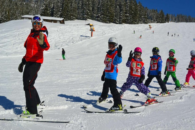 Skilehrerin in roter Jacke mit einer Gruppe von Kindern in bunten Skianzügen auf einer verschneiten Piste.