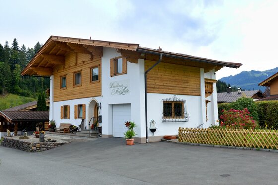 Zweistöckiges Landhaus mit weißer Fassade, Holzverkleidung im Obergeschoss und flachem Satteldach vor Bergkulisse.