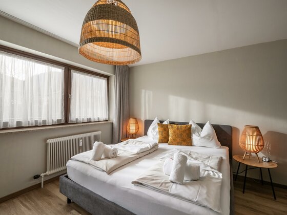 Schlafzimmer Appartement Hafkamp Modernes Doppelbett mit weißer Bettwäsche, gelben Kissen und Rattanlampen in einem hellen Schlafzimmer.
