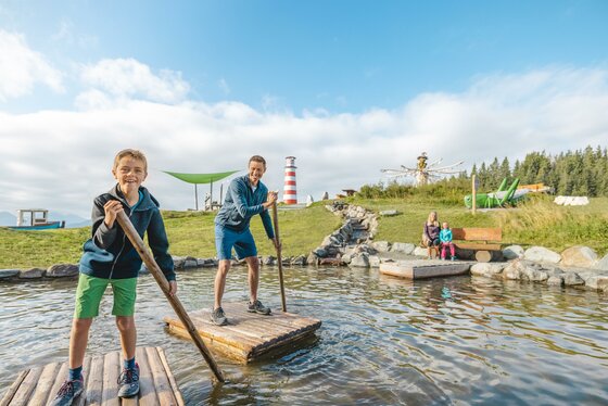 Flossfahrt Zwei Kinder stehen auf einer Holzplattform im Wasser und haben Spaß. Im Hintergrund sind grüne Wiesen und ein Leuchtturm zu sehen.