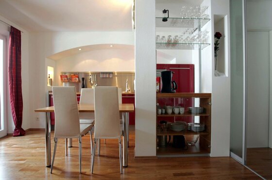 Moderner Essbereich mit Holztisch und weißen Stühlen vor einer offenen Küche mit roten Fronten.