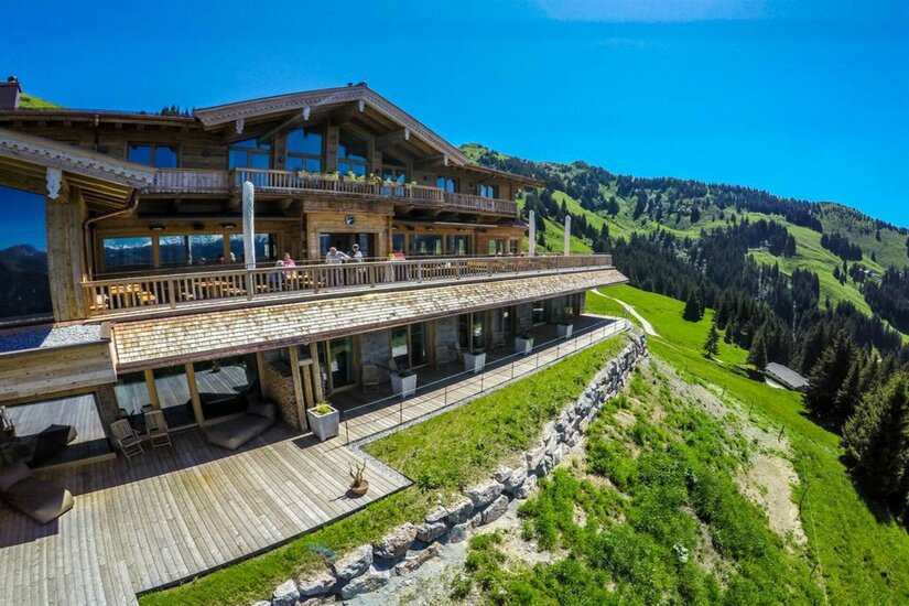 Große Berghütte aus Holz mit Sonnenterasse vor bewaldeten Berghängen unter blauem Himmel.