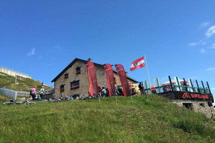 Steinerne Berghütte mit Terrasse, Fahnen und Mountainbikes auf einer grünen Wiese unter blauem Himmel.