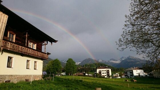 Fichterhof Kitzbühel im Sommer Alpenhaus mit Holzbalkon vor einer Bergkulisse mit Regenbogen unter bewölktem Himmel.