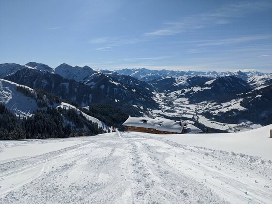 Blick von einer verschneiten Skipiste auf eine Almhütte und die umliegende Berglandschaft im Winter.