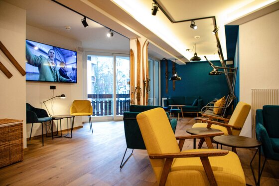 Lounge im Safestay Kitzbühel Moderner Aufenthaltsraum mit gelben und blauen Sesseln, Fernseher und dekorativen Holzskiern an der Wand.