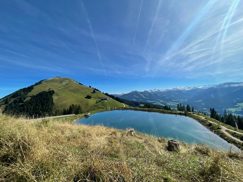 Ein künstlicher Speichersee auf einem Bergrücken vor grasbewachsenen Gipfeln und blauem Himmel.