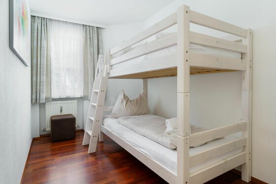 Ein helles Zimmer mit einem Etagenbett aus weißem Holz, Parkettboden und einem Fenster mit Vorhang.